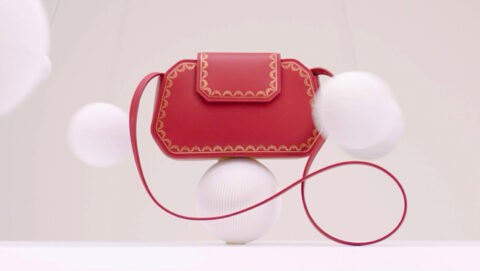 A stylish red handbag with gold trimmings is artistically balanced atop white spherical objects against a soft beige background, evoking a sense of elegance and minimalism.