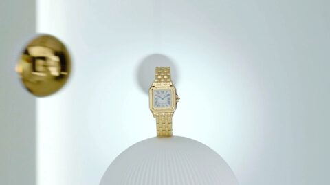 A golden wristwatch with a rectangular face on a white spherical pedestal against a soft white background, showcasing an elegant and minimalist display.