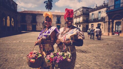 Two women dressed in traditional clothing, smiling and carrying baskets of vibrant flowers, on a sunny day in a historic square with cobblestones and old buildings in the background.