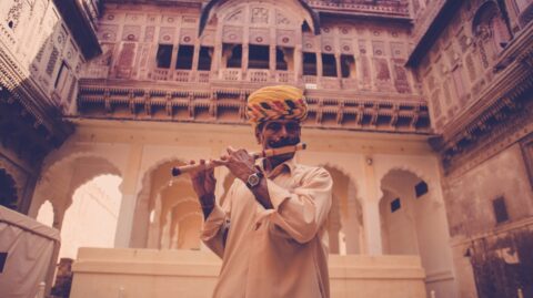 A musician, clad in traditional attire with a vibrant turban, serenades with a flute within the ornate courtyard of an indian palace.