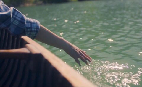 Gentle touch: a person in a checkered shirt resting their hand on the side of a wooden boat, trailing their fingers through the calm, sparkling waters.