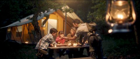 A family enjoying a warm, cozy evening together while camping in the woods, with softly illuminated tents in the background and the glow of a lantern hanging overhead.