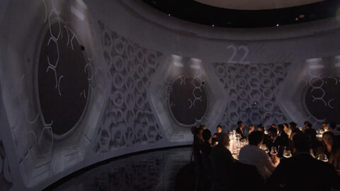 Un groupe de personnes assises autour de tables dans une salle futuriste avec des murs recouverts de motifs géométriques et des écrans circulaires. L'éclairage est tamisé, créant une ambiance immersive.