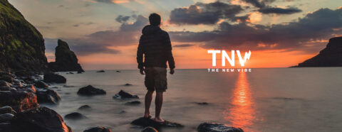 Homme debout sur des rochers au bord de la mer, regardant un coucher de soleil avec le texte "TNV THE NEW VIBE".