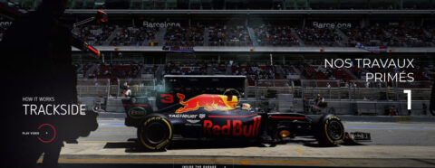 L'image montre une scène de piste de course automobile avec une voiture de Formule 1 de l'équipe Red Bull Racing au centre. L'arrière-plan montre des tribunes remplies de spectateurs avec l'inscription "Barcelona" visible. Le texte "HOW IT WORKS TRACKSIDE" est affiché à gauche avec une option pour "PLAY VIDEO". À droite, le texte "NOS TRAVAUX PRIMÉS 1" est affiché, suggérant que cette image fait partie d'une série mettant en avant des réalisations ou des travaux récompensés.