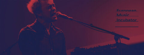 Chanteur jouant du clavier sur scène avec l'inscription "European Music Incubator" à droite.