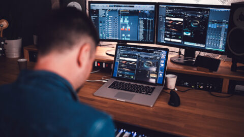L'image montre une personne travaillant sur un ordinateur portable et deux écrans de moniteur dans un studio de musique ou un environnement de production sonore. Les écrans affichent des logiciels de production musicale, avec diverses interfaces de mixage et de création de sons visibles. La personne semble être concentrée sur son travail, peut-être en train de composer ou de mixer de la musique. L'arrière-plan comporte des équipements audio supplémentaires, comme des enceintes et d'autres instruments de musique, suggérant un espace bien équipé pour la production sonore.